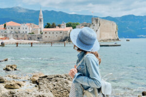 Back view of curly hair woman in hat standing in blue dress looking out towards blue sea and sky in Montenegro.